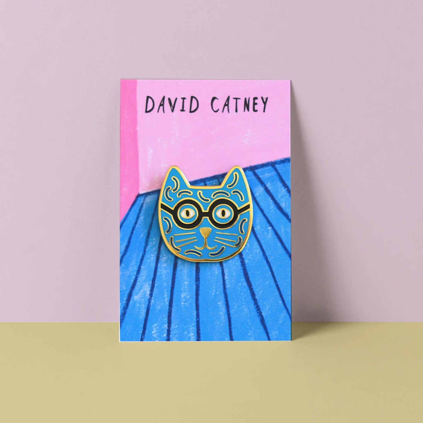 Enamel pin of a cat with glasses on a blue and pink card with 'David Catney' branding.