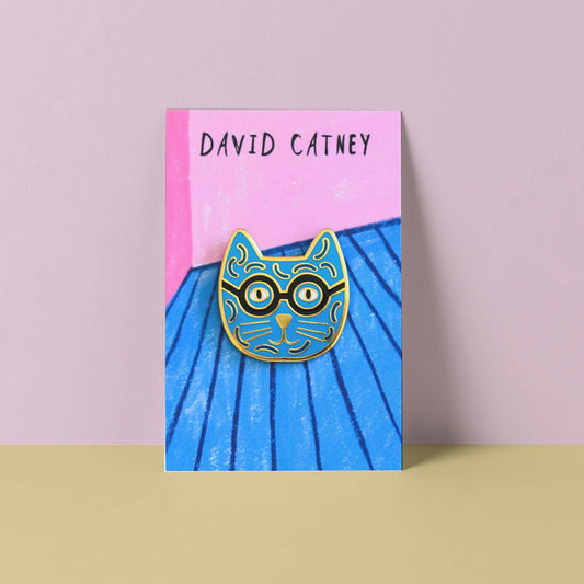 Enamel pin of a cat with glasses on a blue and pink card with 'David Catney' branding.